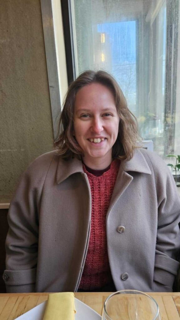 Lilli, a white woman, with shoulder length blond hair, smiles sitting at a sunny restaurant table. She wears a tan coat over a red sweater, and her eyes crinkle up over her broad and crooked-toothed grin.